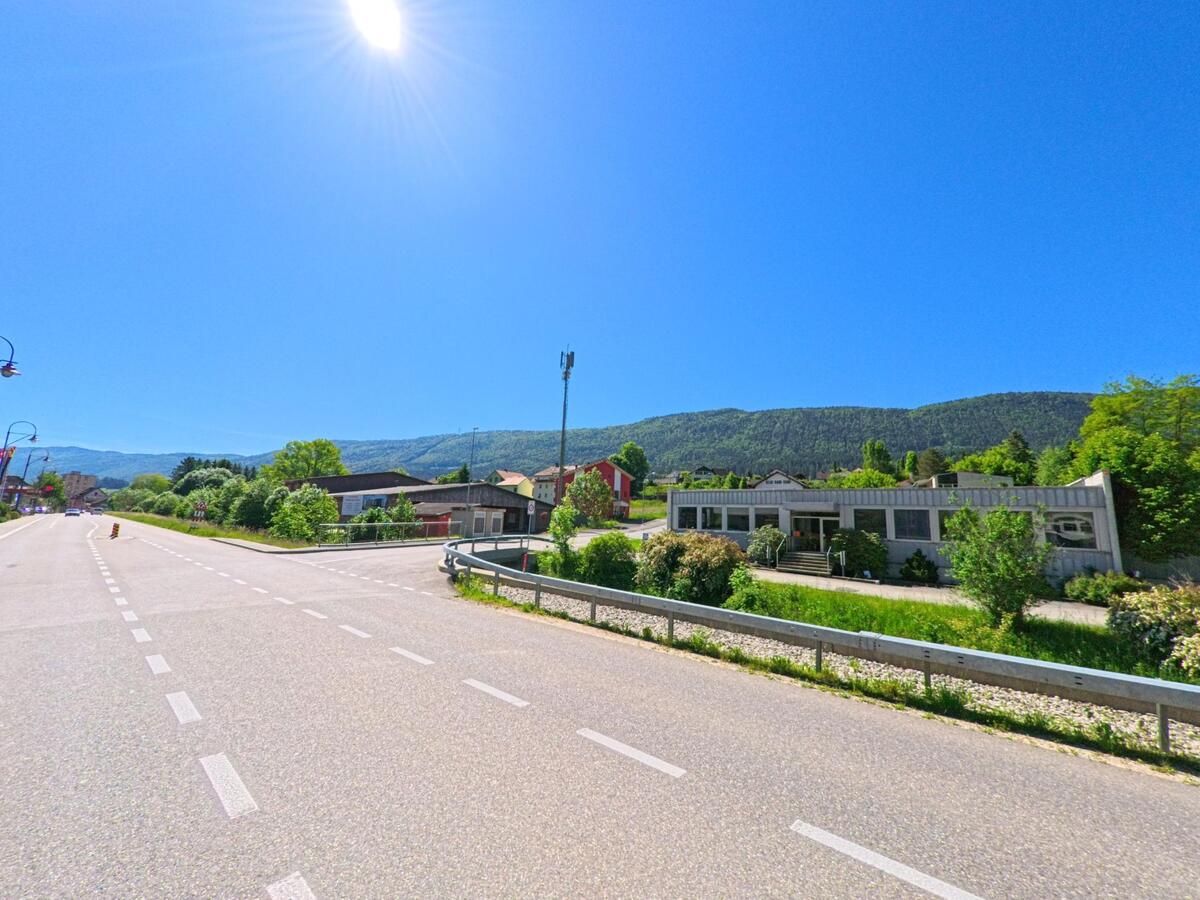 Entlang der Hauptstrasse von Champoz (2735, Kanton Bern) liegen mehrere Gebaeude und ein modernes Flachdachhaus vor dem bewaldeten Jurahang unter strahlender Sonne.