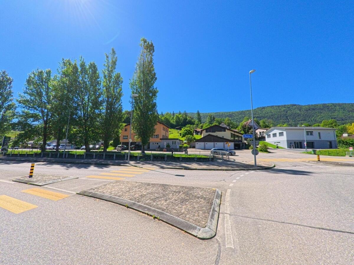 An einer breiten Kreuzung in Champoz (2735, Kanton Bern) stehen hohe Baeume und Wohnhaeuser vor dem bewaldeten Hang, waehrend gelbe Markierungen den Fussgaengeruebergang hervorheben.