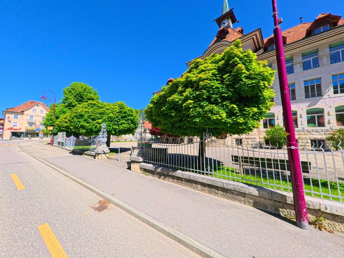 Vor dem markanten Schulgebaeude von Champoz (2735, Kanton Bern) stehen dichte, rund geschnittene Baeume hinter einem Metallzaun entlang der ruhig verlaufenden Strasse.