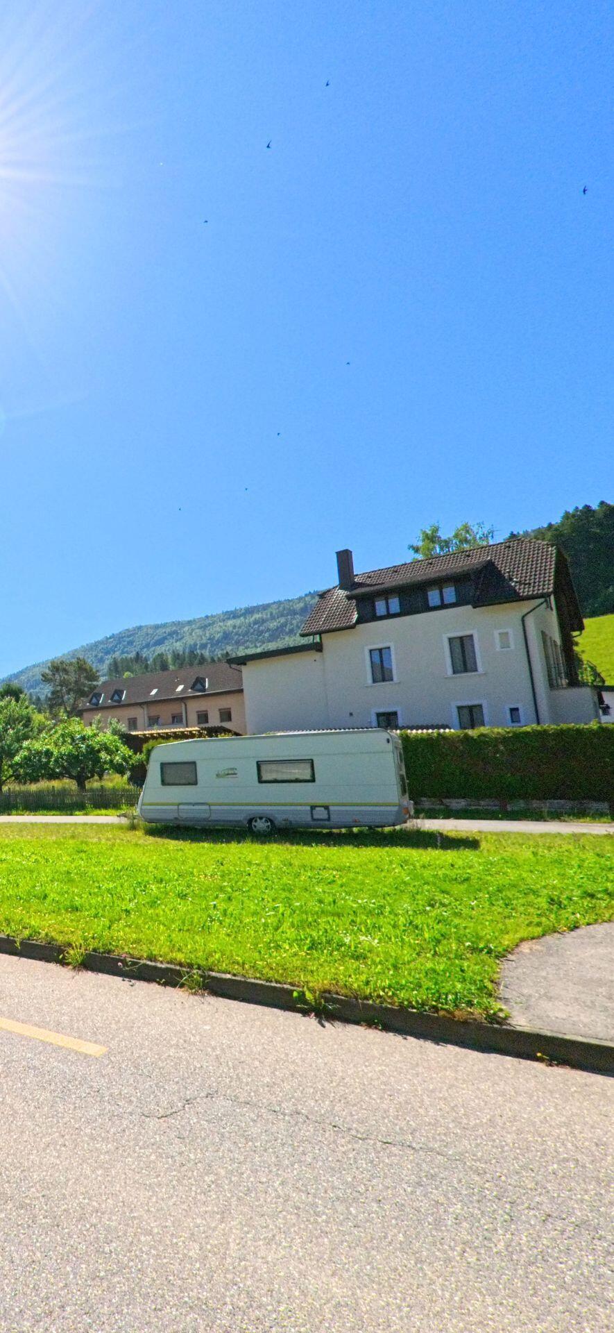 Auf einer gruene Flaeche am Siedlungsrand von Champoz (2735, Kanton Bern) steht ein Wohnwagen vor einem hellen Mehrfamilienhaus mit Blick auf den bewaldeten Hang.