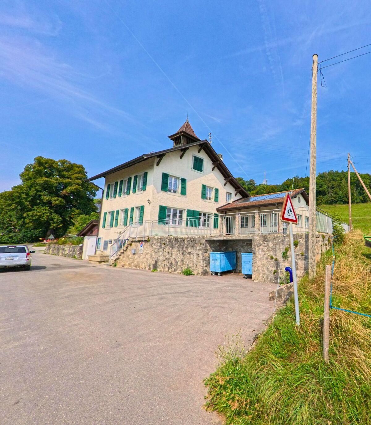 Ein helles Haus mit grünen Fensterläden steht in Chardonne (1801, Kanton Waadt) am Rand einer Strasse mit Blick auf Wiesen und Wald.