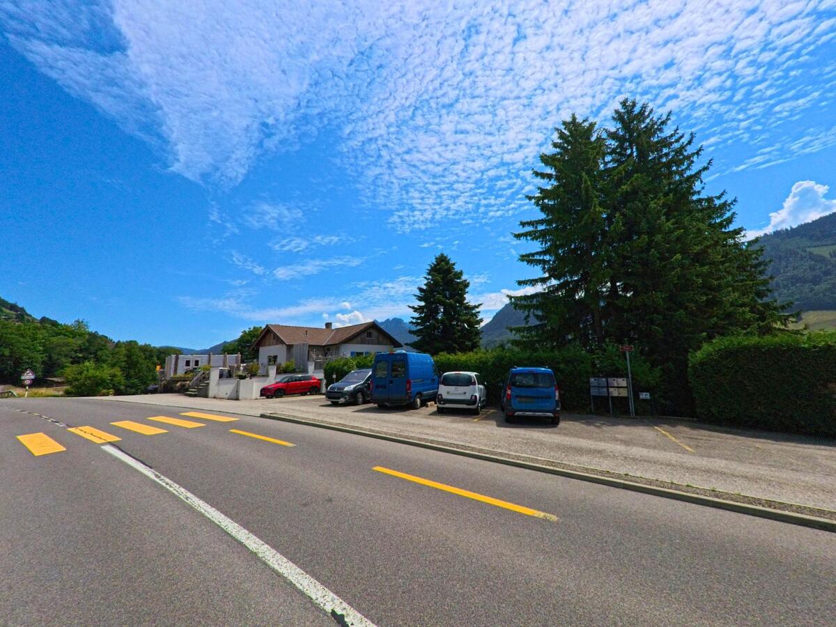 An einer Strasse in Châtel-sur-Montsalvens (1653, Kanton Freiburg) liegen mehrere geparkte Autos neben Wohnhäusern und hohen Tannen unter blauem Himmel.