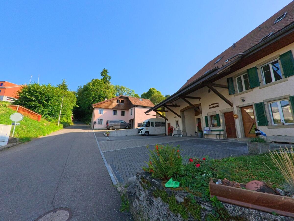 In Châtillon (1473, Kanton Freiburg) fuehrt eine ansteigende Strasse an traditionellen Wohnhaeusern mit gruenen Fensterlaeden vorbei, waehrend im Hintergrund Baeume und moderne Gebaeude zu sehen sind.