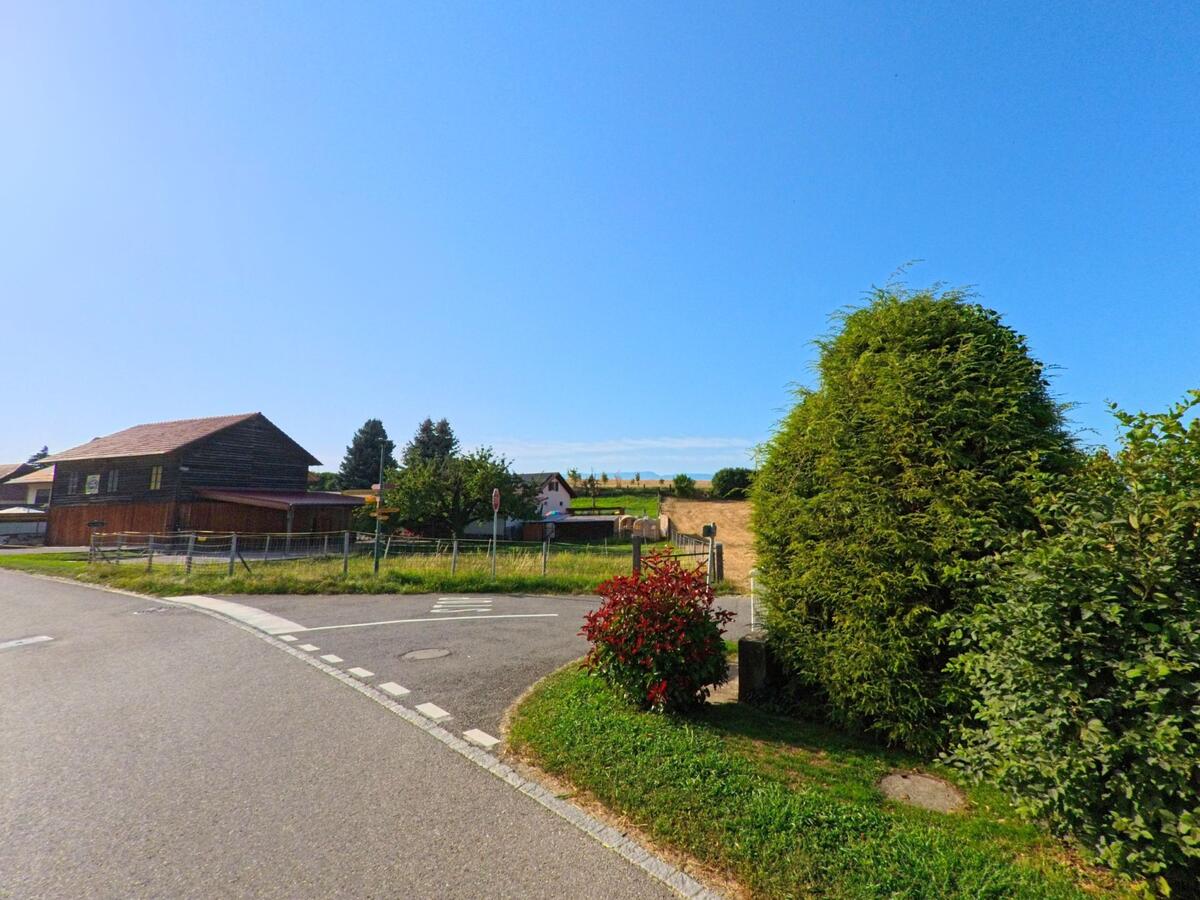 In Châtillon (1473, Kanton Freiburg) fuehrt eine Strasse an einer Hofanlage mit Holzgebaeude vorbei, waehrend im Hintergrund Felder und weitere Haeuser liegen.