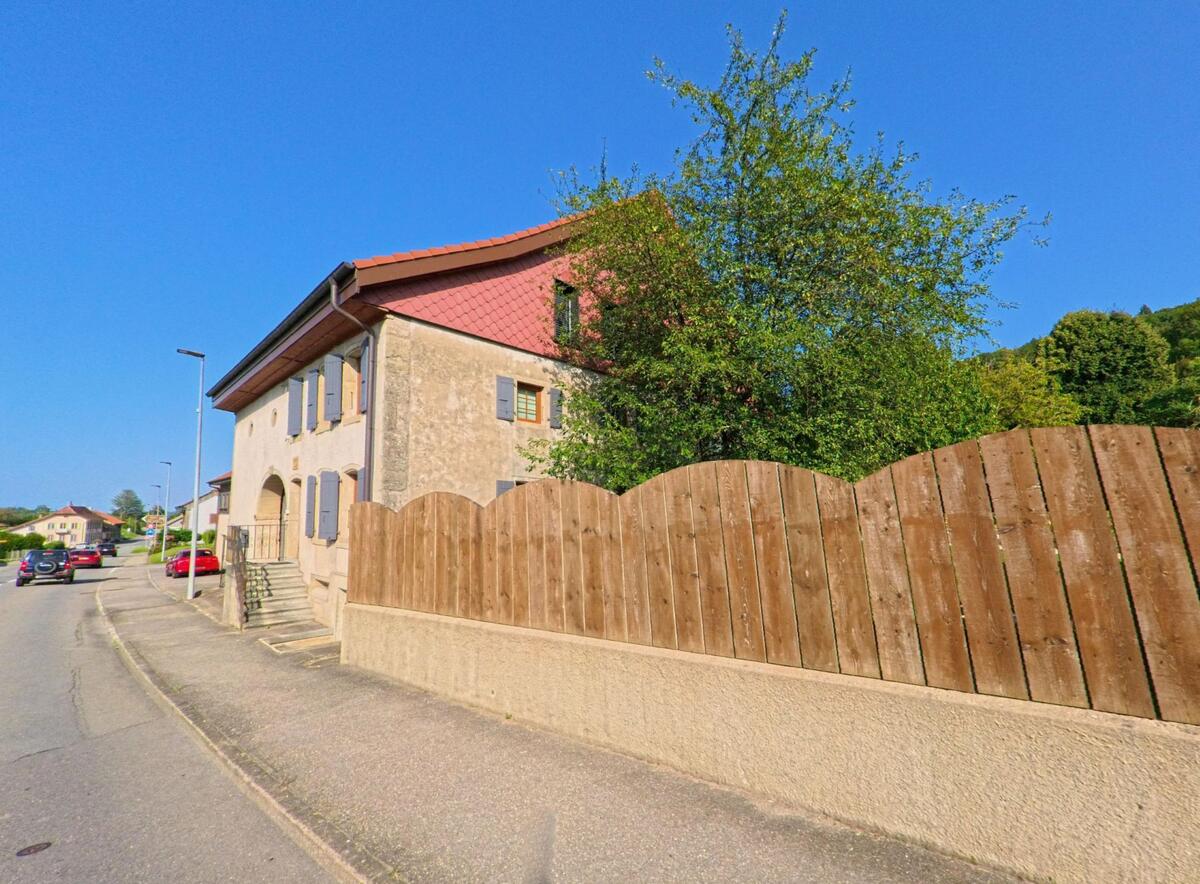 In Châtillon (1473, Kanton Freiburg) steht ein traditionelles Wohnhaus mit steinernem Torbogen und blaugrauen Fensterlaeden an einer Strasse, die von mehreren Autos befahren wird.