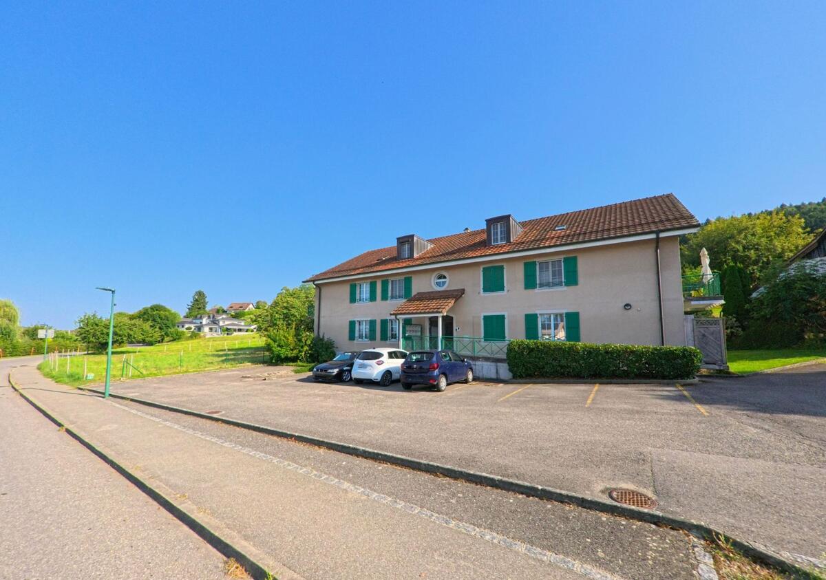 In Châtillon (1473, Kanton Freiburg) steht ein langgestrecktes Wohnhaus mit gruenen Fensterlaeden und vorgelagertem Parkplatz am Rand einer Strasse.