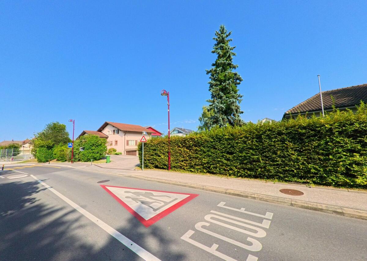 Auf der Fahrbahn in Chavannes-de-Bogis (1279, Kanton Waadt) weist die Markierung „Ecole“ mit Piktogramm auf einen nahen Schulbereich hin.