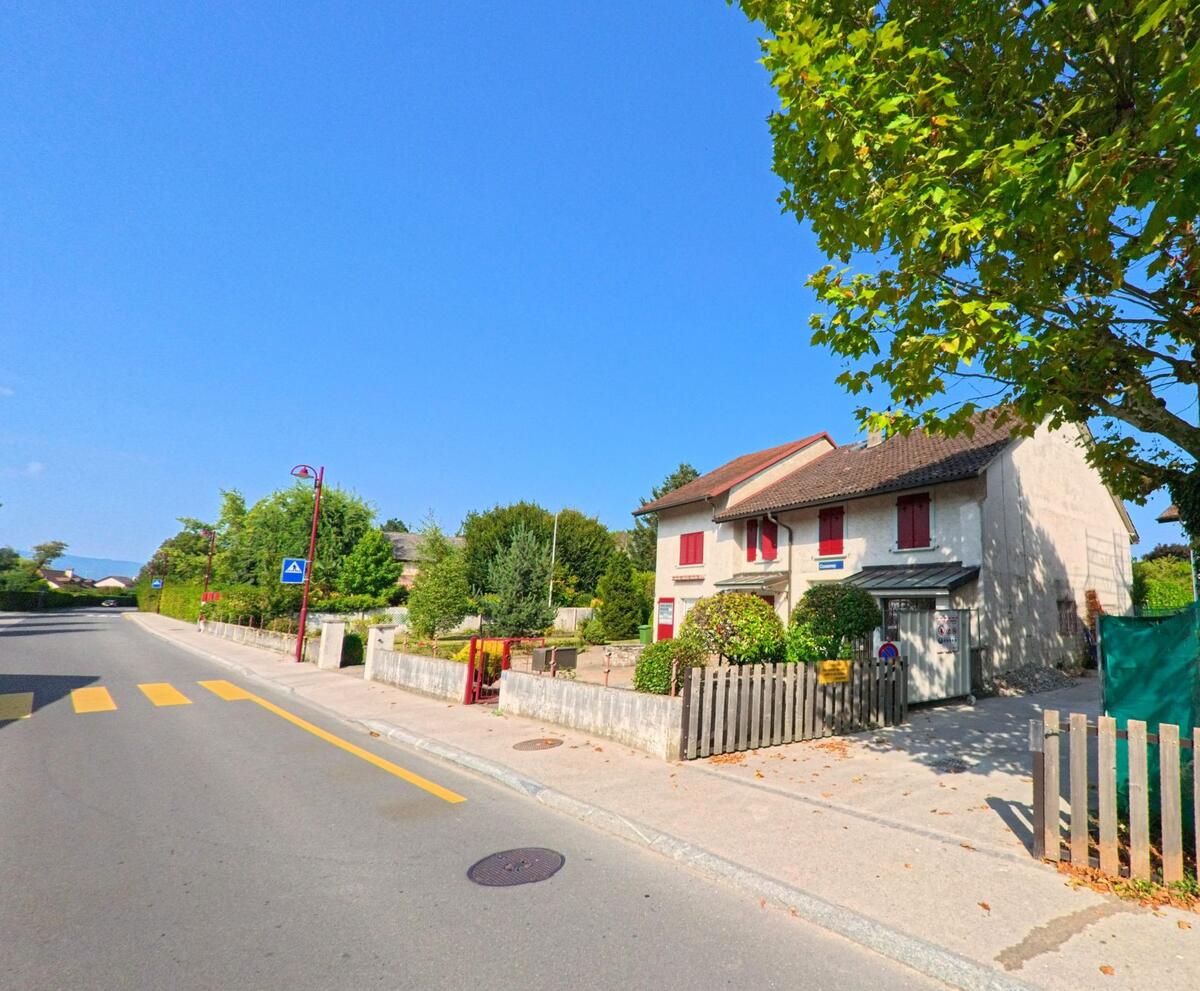Ein Wohnhaus mit roten Fensterläden und Vorgarten steht an einer Strasse mit Zebrastreifen in Chavannes-de-Bogis (1279, Kanton Waadt).
