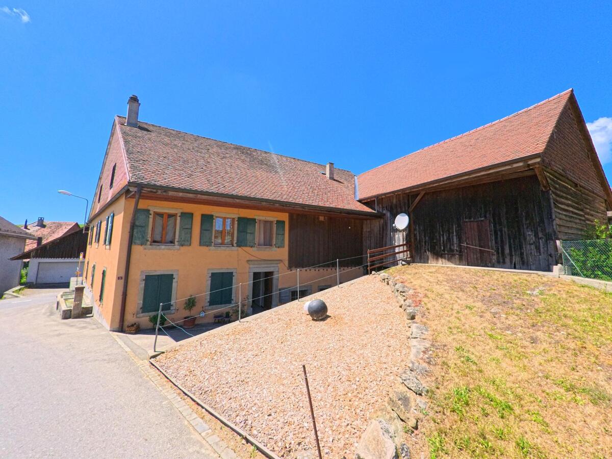 In Chavannes-sur-Moudon (1512, Kanton Waadt) stehen ein traditionelles Wohnhaus mit grünen Fensterläden und eine angrenzende Scheune mit Holzwandseite an einem Hanggrundstück.