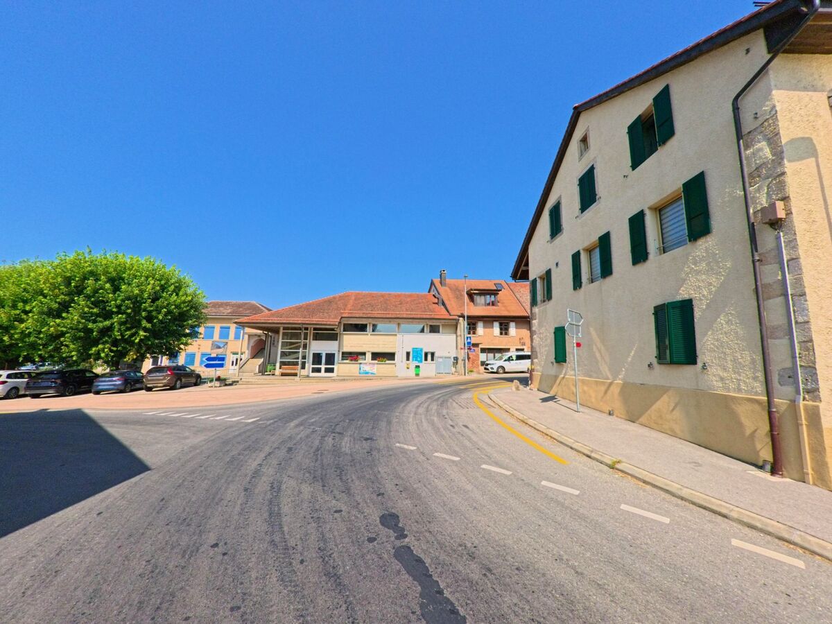 Eine sanft gekruemmte Strasse fuehrt im Zentrum von Chavannes-le-Veyron (1148, Kanton Waadt) an hellen Gebaeuden mit gruenen Fensterlaeden und einem modernen Gemeindesaal vorbei.