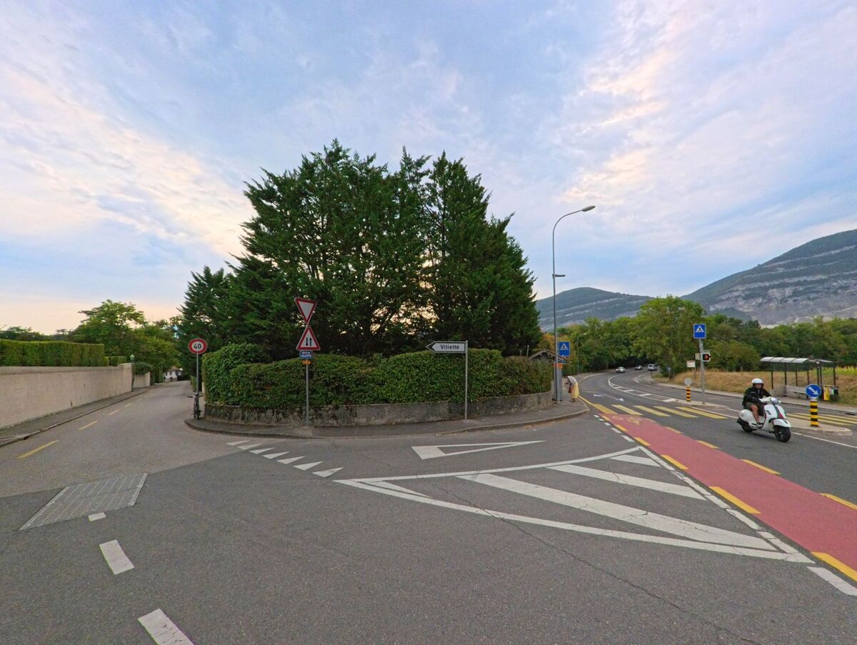 Eine Kreuzung in Chene-Bougeries (1224, Kanton Genf) mit roten Radstreifen fuehrt in Richtung Villette, waehrend ein Motorroller die Strasse entlangfaehrt und im Hintergrund Huegel sichtbar sind.