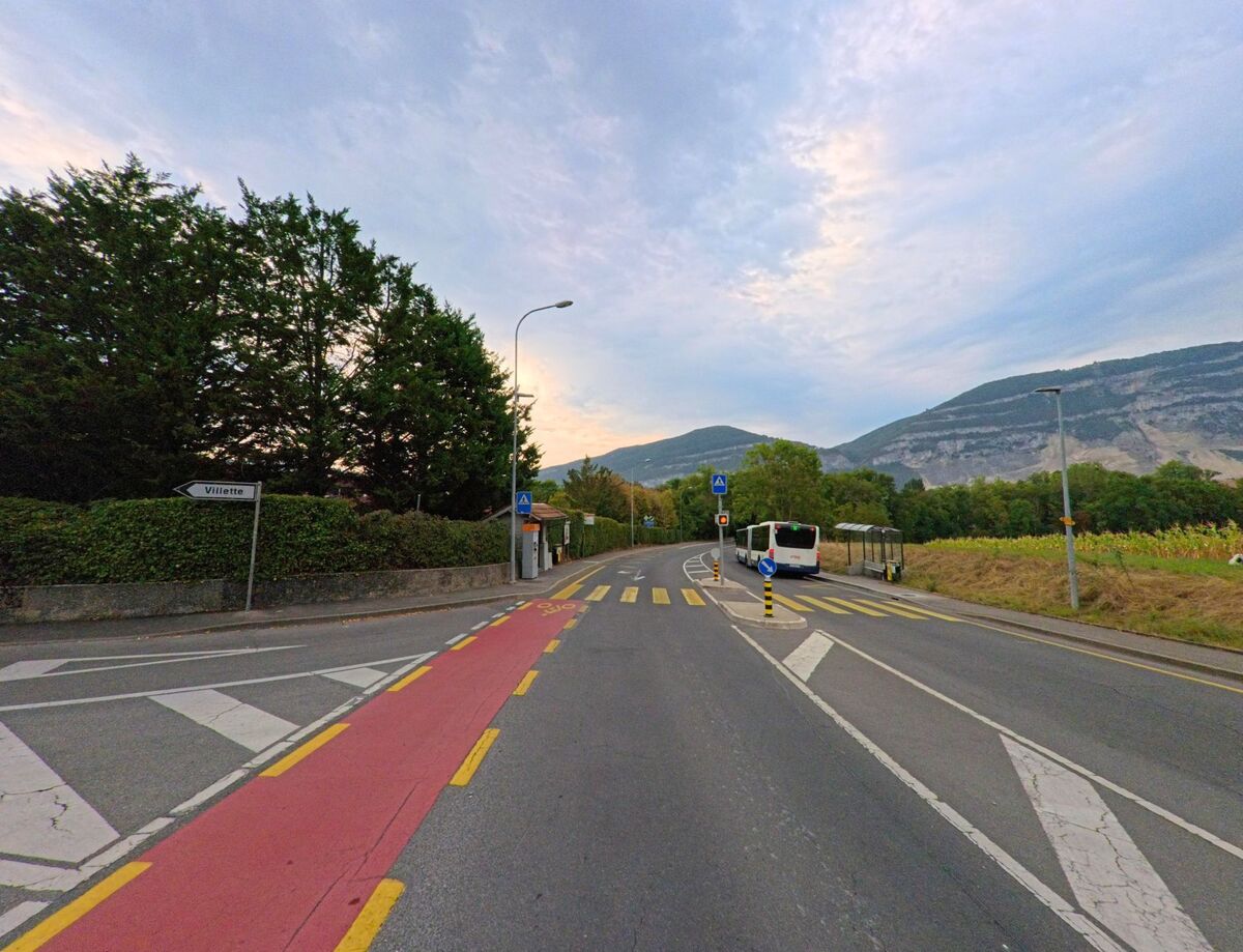 Eine Landstrasse in Chene-Bougeries (1224, Kanton Genf) fuehrt an einer Bushaltestelle vorbei, im Hintergrund erheben sich bewaldete Huegel und ein Bus haelt am Fussgaengerstreifen.