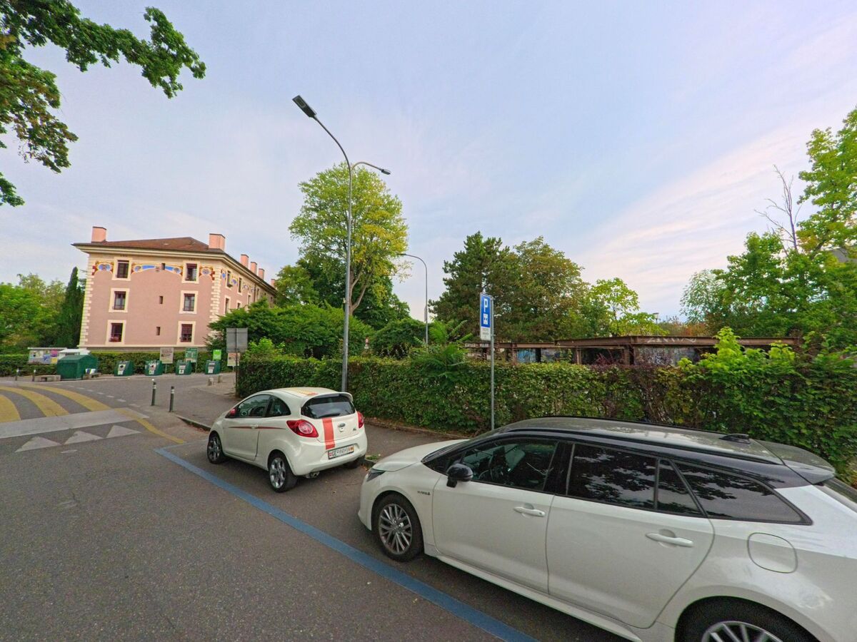 Ein rosa gestrichenes Mehrfamilienhaus in Chene-Bougeries (1224, Kanton Genf) mit dekorativen Malereien steht nahe einer Strasse, davor befinden sich Parkplaetze und Container.