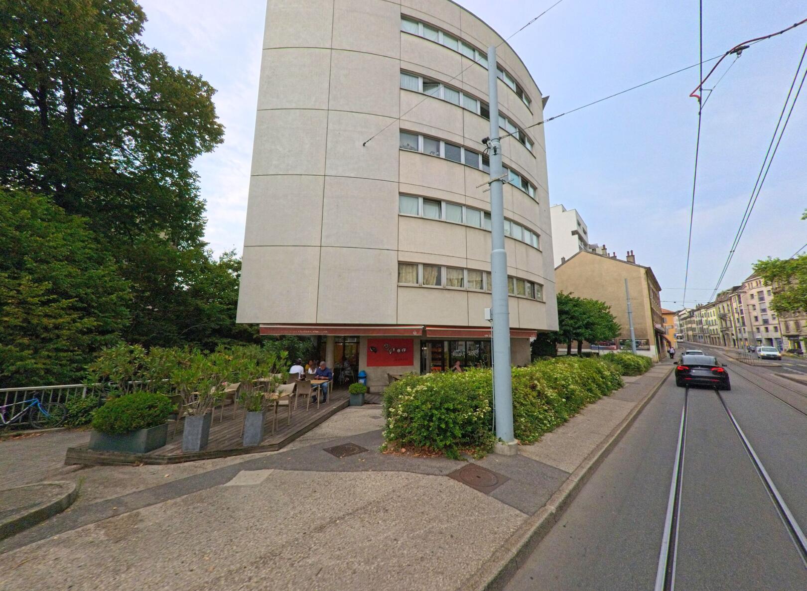 An einer Strasse in Chene-Bougeries (1224, Kanton Genf) steht ein modernes Wohnhaus mit abgerundeter Fassade, im Erdgeschoss befindet sich ein Restaurant mit Aussenbestuhlung.