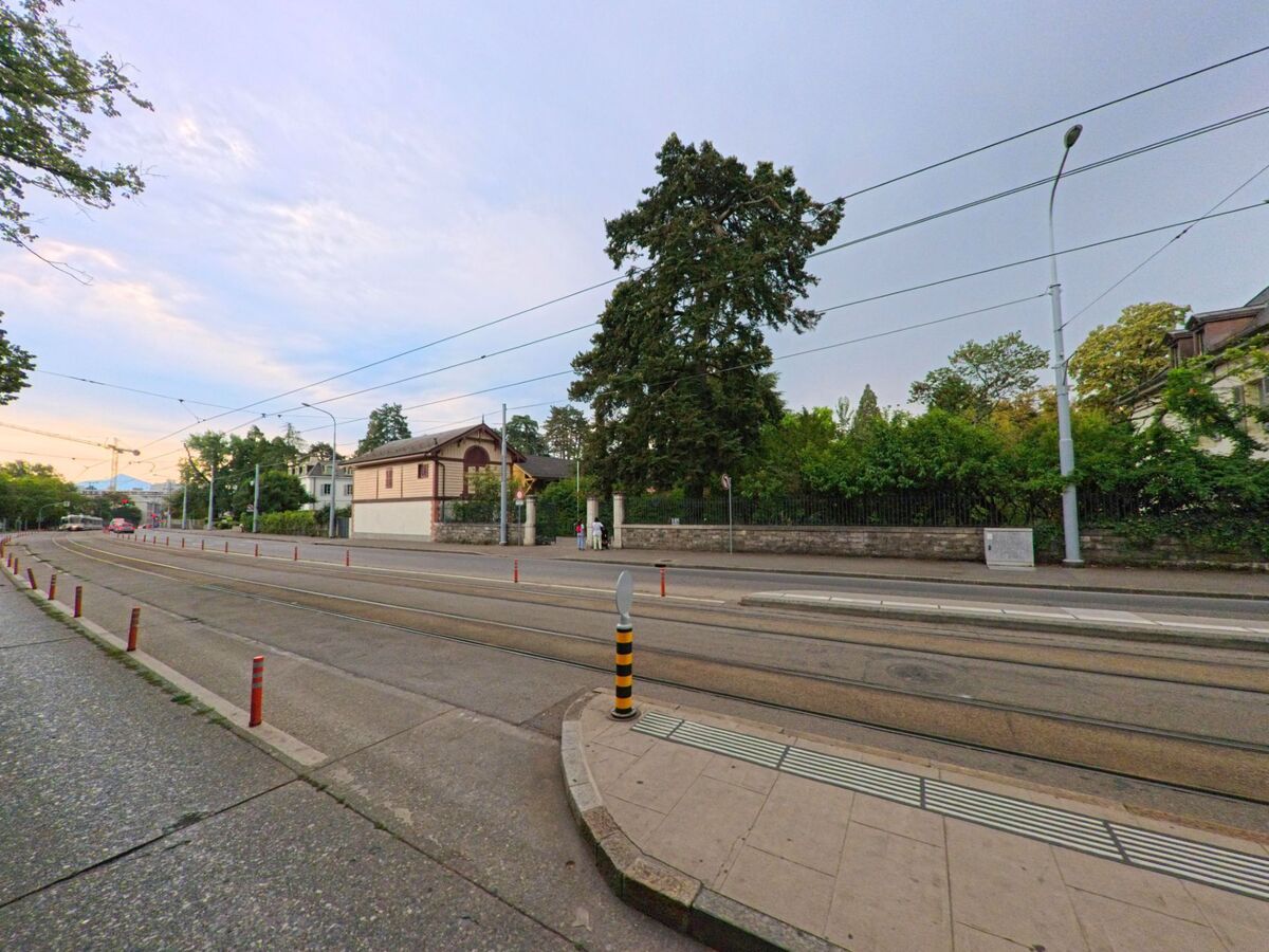 Eine Strassenbahntrasse verläuft durch Chene-Bougeries (1224, Kanton Genf), daneben stehen ein kleines Gebaeude mit Holzfassade und grosse Baeume hinter einer Steinmauer.