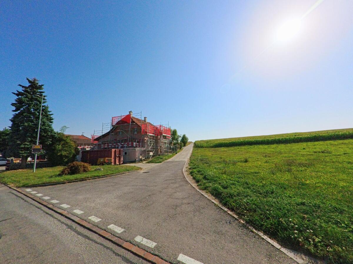 Ein Haus mit Baugerüst steht am Dorfrand von Chêne-Pâquier (1464, Kanton Waadt) neben einer langen Strasse durch grüne Felder.