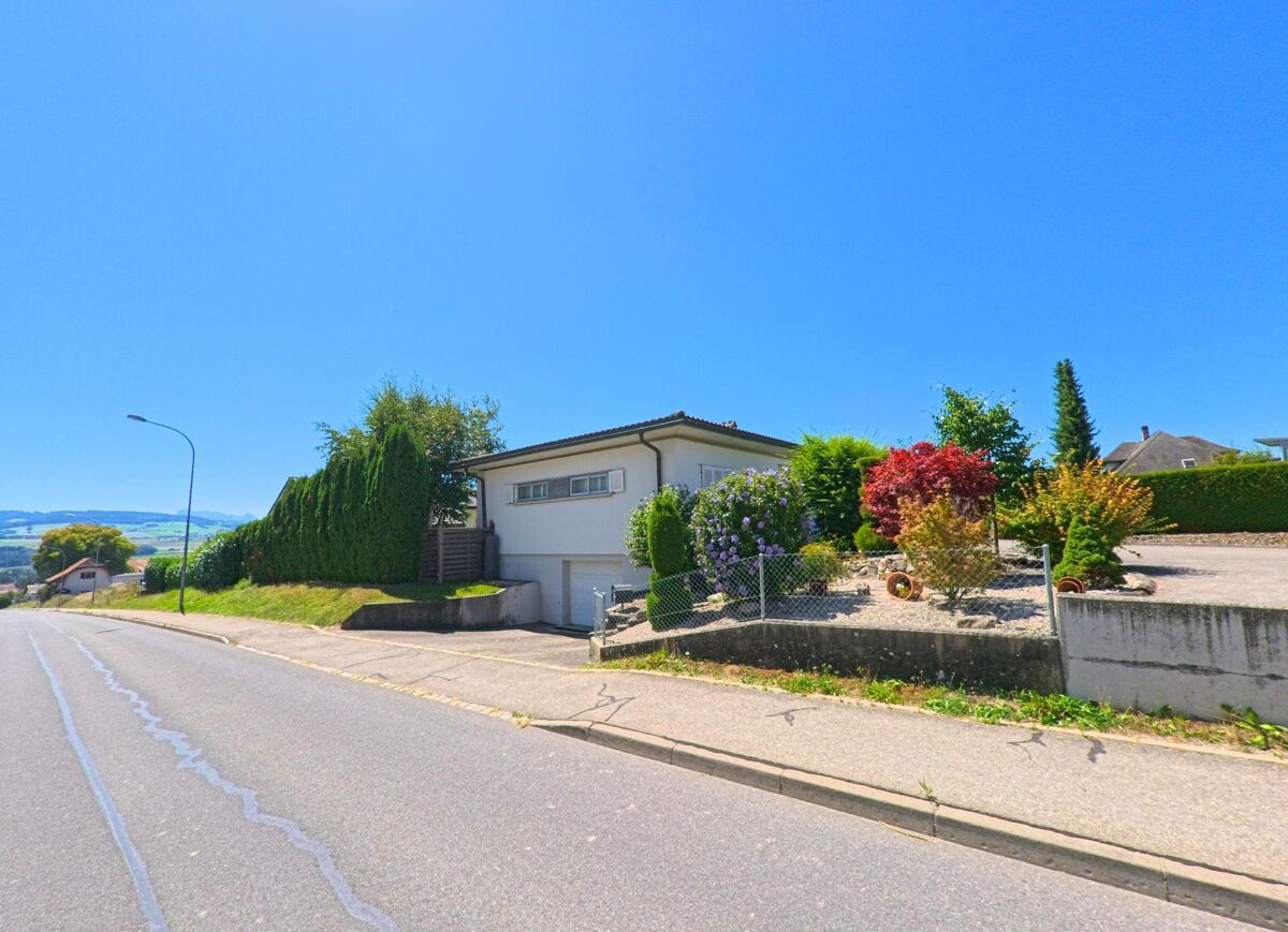 An einer breiten Strasse in Chénens (1744, Kanton Freiburg) steht ein Einfamilienhaus mit gepflegtem Vorgarten, in dem verschiedene Ziersträucher wachsen.