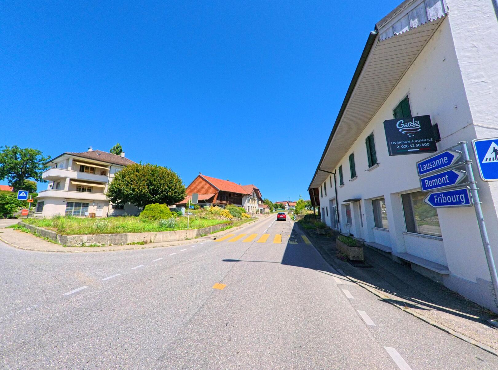 An einer leicht ansteigenden Hauptstrasse in Chénens (1744, Kanton Freiburg) stehen Wohnhäuser und ein Gasthaus, während Wegweiser in Richtung Lausanne, Romont und Fribourg zeigen.