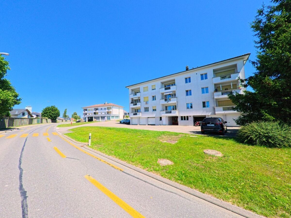An einer sanft gebogenen Strasse in Chénens (1744, Kanton Freiburg) stehen zwei weisse Mehrfamilienhäuser mit Balkonen und Garagenzufahrten.