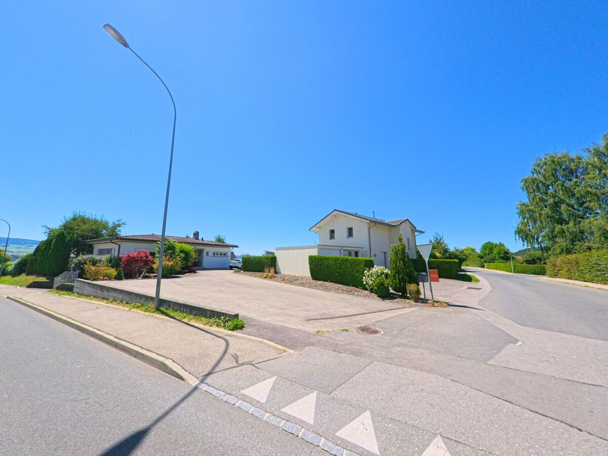 An einer Strassenkreuzung in Chénens (1744, Kanton Freiburg) stehen zwei Einfamilienhäuser mit gepflegten Hecken und Vorgärten.