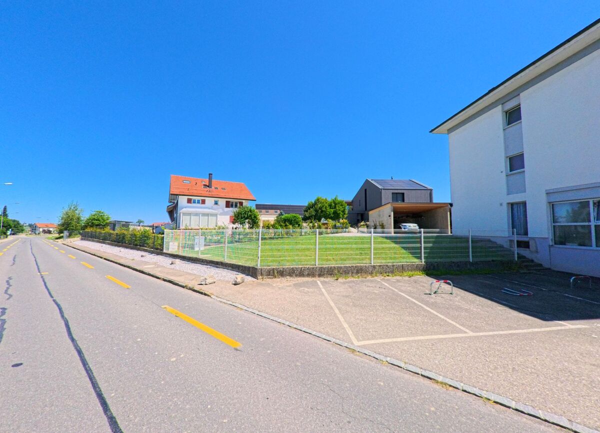 Entlang einer breiten Strasse in Chénens (1744, Kanton Freiburg) stehen mehrere Wohnhäuser mit gepflegten Gärten und Carports.