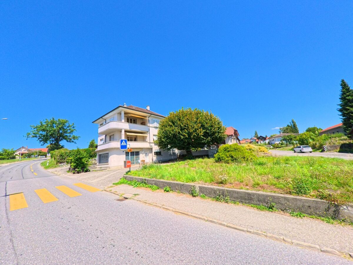 An einer Strassenkurve in Chénens (1744, Kanton Freiburg) steht ein mehrstöckiges Wohnhaus mit Balkonen neben einem Fussgängerstreifen.