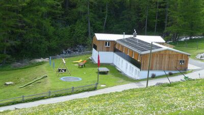 In Madulain (7523, Kanton Graubuenden) liegt die Chesa Algrezia mit Holzfassade und Solarpanelen auf dem Dach, umgeben von Wiese, Spielplatz und Waldrand.
