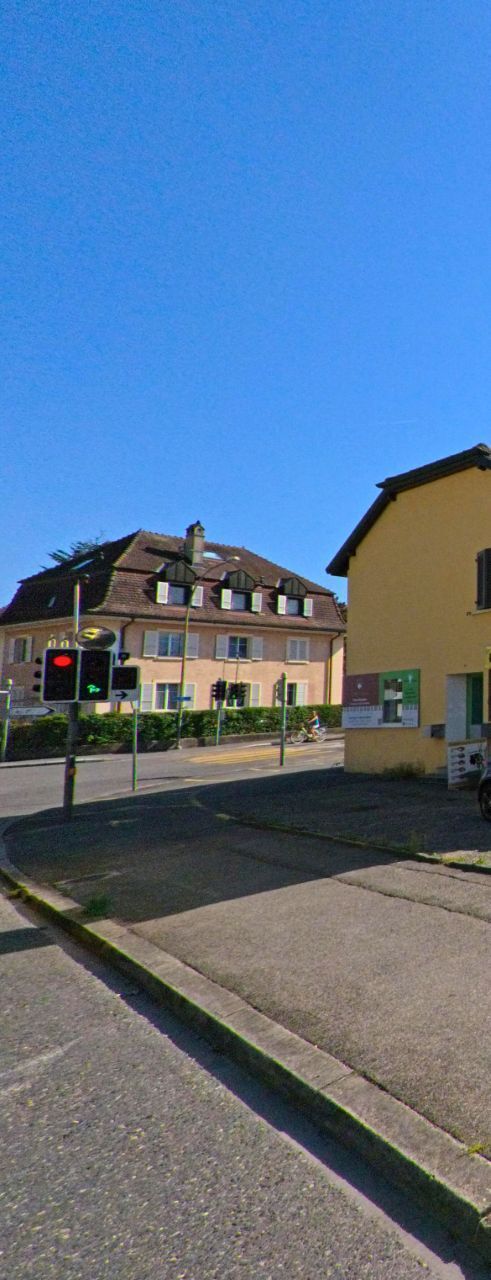 An einer Kreuzung in Cheseaux-Noréaz (1400, Kanton Waadt) steht ein gelbes Gebäude neben einer Verkehrsampel, im Hintergrund sind weitere Wohnhäuser sichtbar.