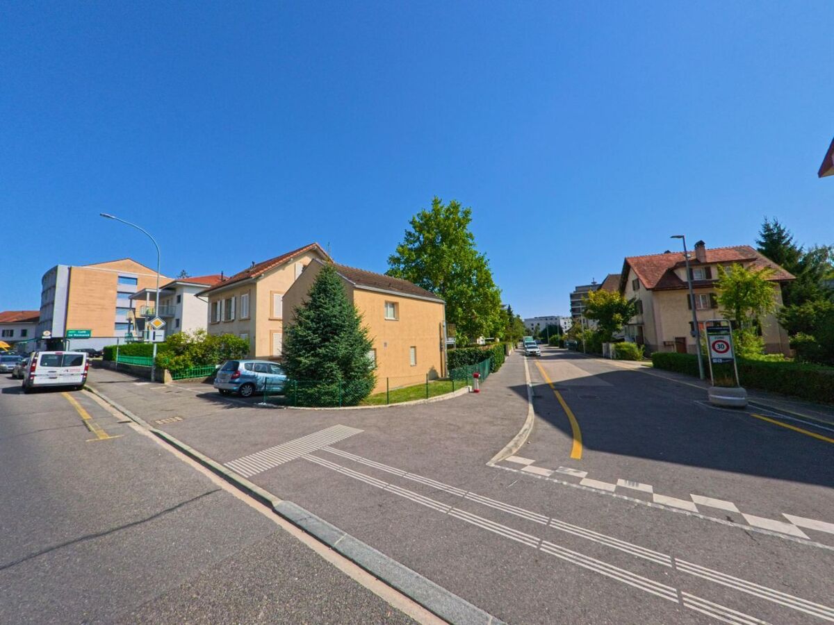 Wohnhäuser und Bäume säumen eine verkehrsberuhigte Strasse in Cheseaux-Noréaz (1400, Kanton Waadt) bei strahlend blauem Himmel.