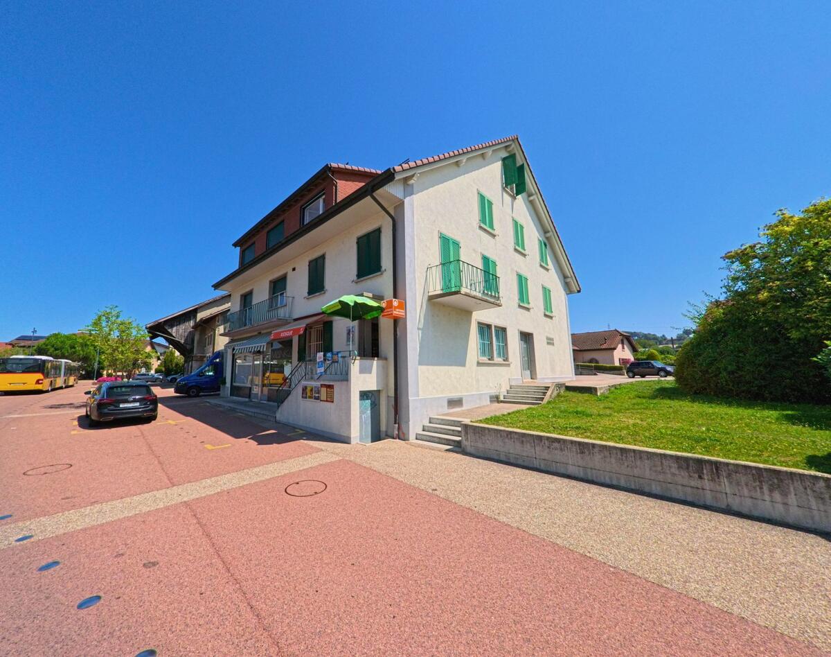 Ein Gebaeude mit gruenen Fensterlaeden in Cheseaux-sur-Lausanne (1033, Kanton Waadt) beherbergt ein kleines Geschaeft, vor dem ein Postauto und mehrere Fahrzeuge stehen.