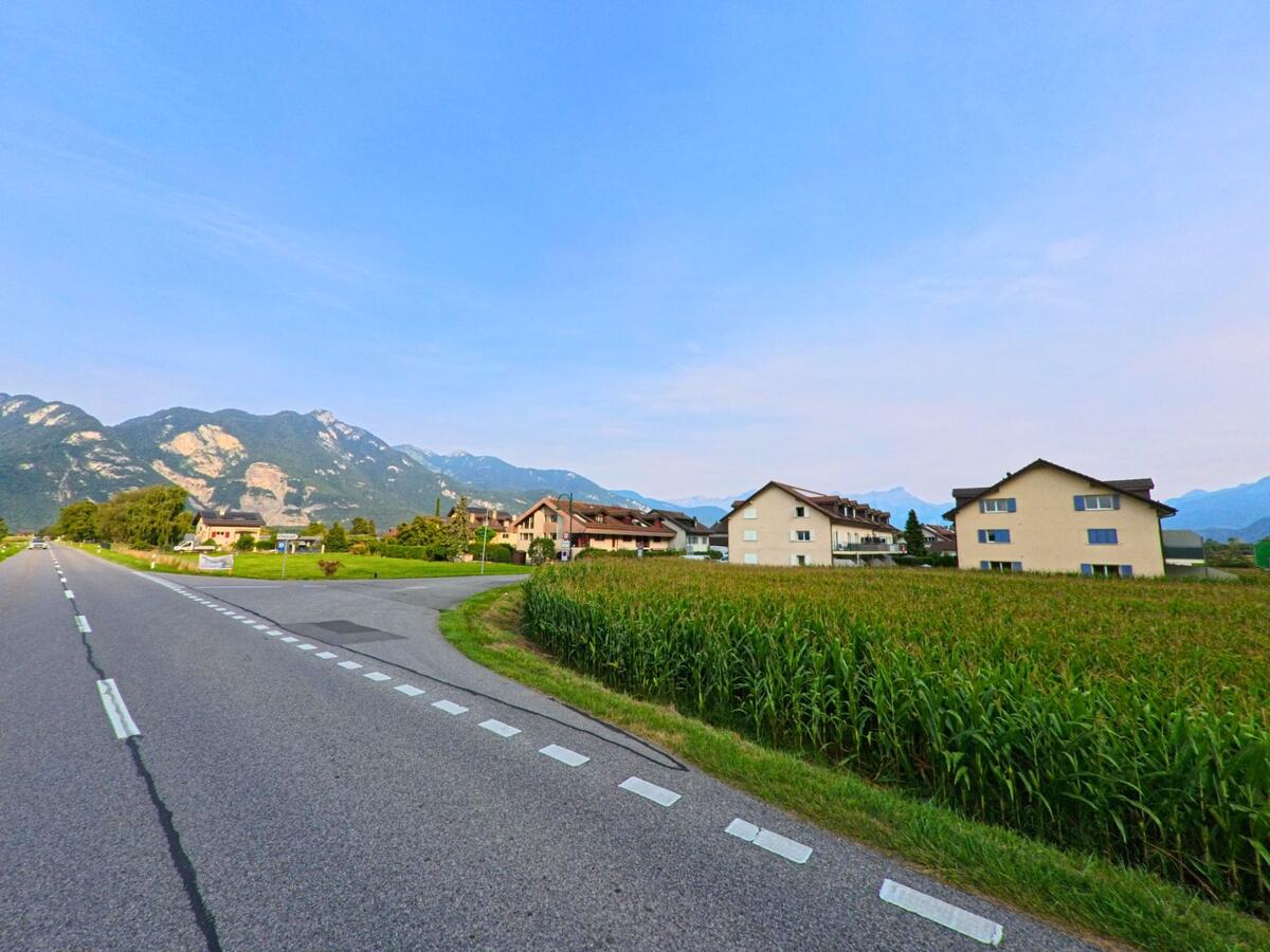 Von der Hauptstrasse in Chessel (1846, Kanton Waadt) blickt man ueber Felder auf mehrere Wohnhaeuser mit den Bergen im Hintergrund.