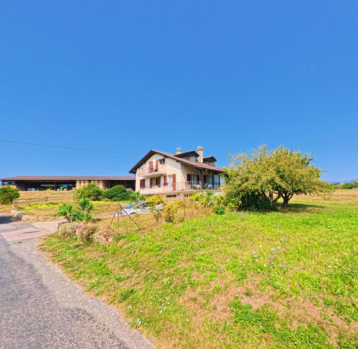 Ein Einfamilienhaus mit Balkon und roten Fensterlaeden liegt in Chevilly (1316, Kanton Waadt) am Rand einer Wiese mit Obstbaum.