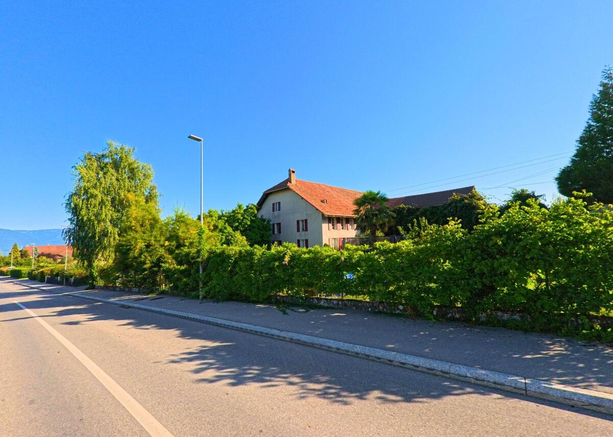 Hinter einer dichten grünen Hecke steht in Chevroux (1545, Kanton Waadt) ein Wohnhaus mit roten Fensterläden und grossem Satteldach an einer breiten Strasse.