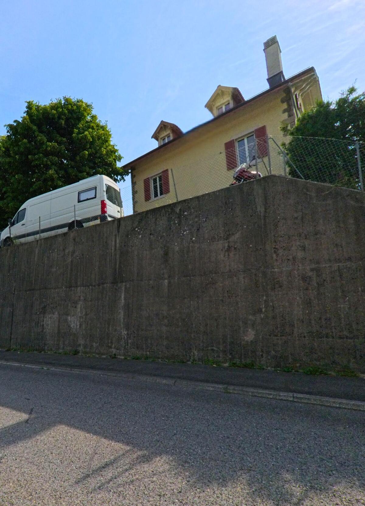 Ein helles Haus mit roten Fensterläden steht in Chexbres (1071, Kanton Waadt) oberhalb einer hohen Betonmauer, neben einem geparkten Lieferwagen.