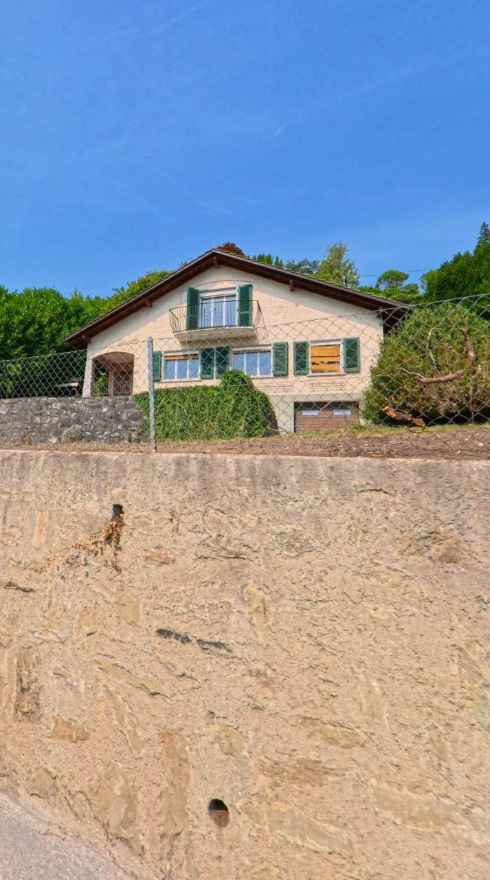 Ein Wohnhaus mit grünen Fensterläden und kleinem Balkon steht in Chexbres (1071, Kanton Waadt) hinter einer Steinmauer unter blauem Himmel.