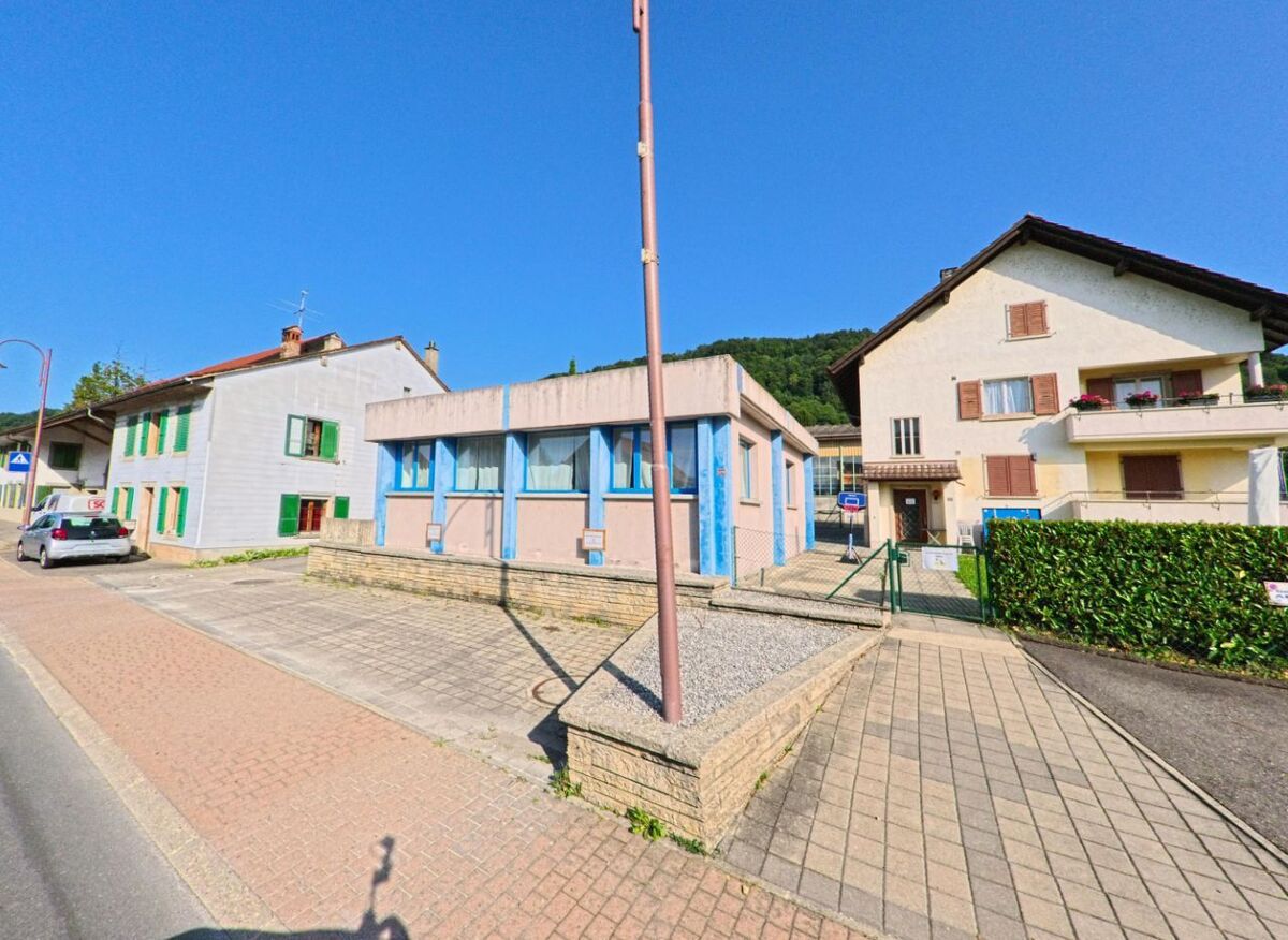 Ein flaches Gebaeude mit blauen Fensterrahmen steht in Cheyres-Chables (1468, Kanton Freiburg) zwischen traditionellen Wohnhaeusern mit Balkonen.