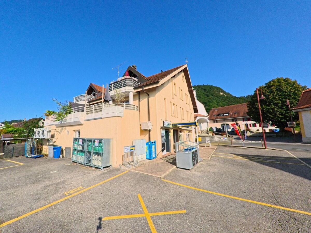 Auf einem Parkplatz in Cheyres-Chables (1468, Kanton Freiburg) steht ein helles Gebaeude mit Geschaeftseingang und mehreren Balkonen im Obergeschoss.