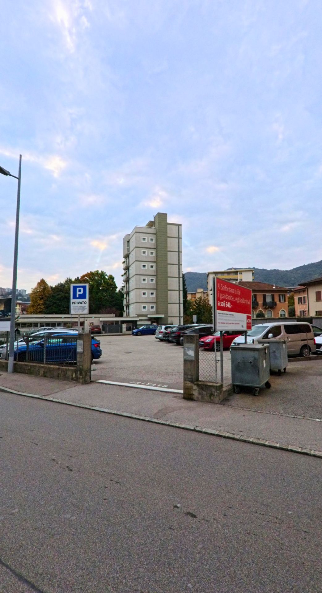 Ein Parkplatz in Chiasso (6832, Kanton Tessin) mit mehreren geparkten Autos liegt vor einem modernen Wohnhaus und umgebenden Gebaeuden am Fusse der Huegel.