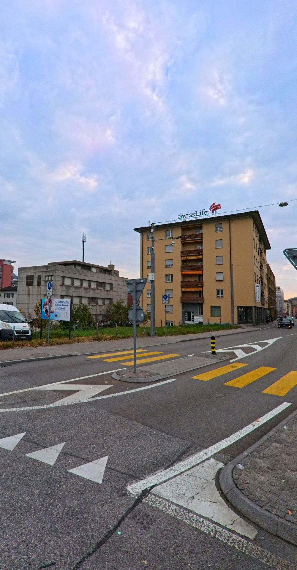 Ein Strassenkreuz in Chiasso (6832, Kanton Tessin) mit einem markanten Gebaeude der Swiss Life und einem Fussgaengerstreifen unter bedecktem Himmel.