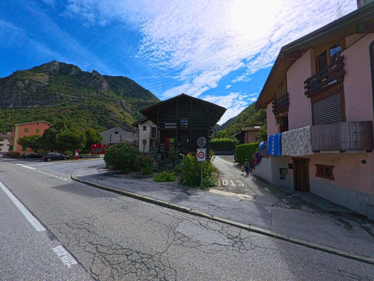 Traditionelle Wohnhaeuser in Chippis (3965, Kanton Wallis) stehen am Strassenrand, im Hintergrund ragen die bewaldeten Berge auf.