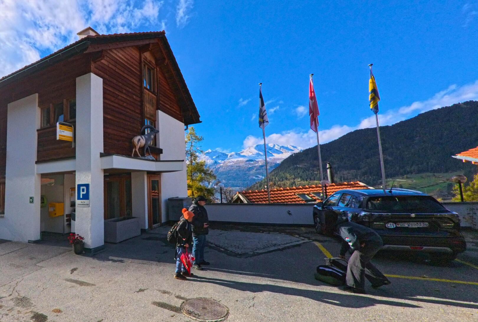 Vor einem Gebäude in Churwalden (7075, Kanton Graubünden) stehen mehrere Personen auf einem Parkplatz, während im Hintergrund Berge und Fahnen unter blauem Himmel zu sehen sind.