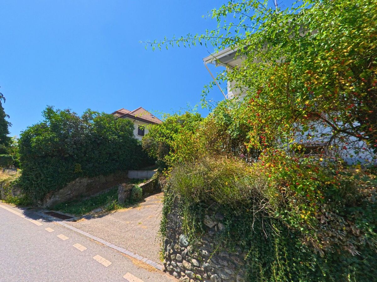 Ein schmaler Aufgang mit Natursteinmauer und dichter Begrünung führt in Clarmont (1127, Kanton Waadt) zu einem Wohnhaus mit rotem Ziegeldach.