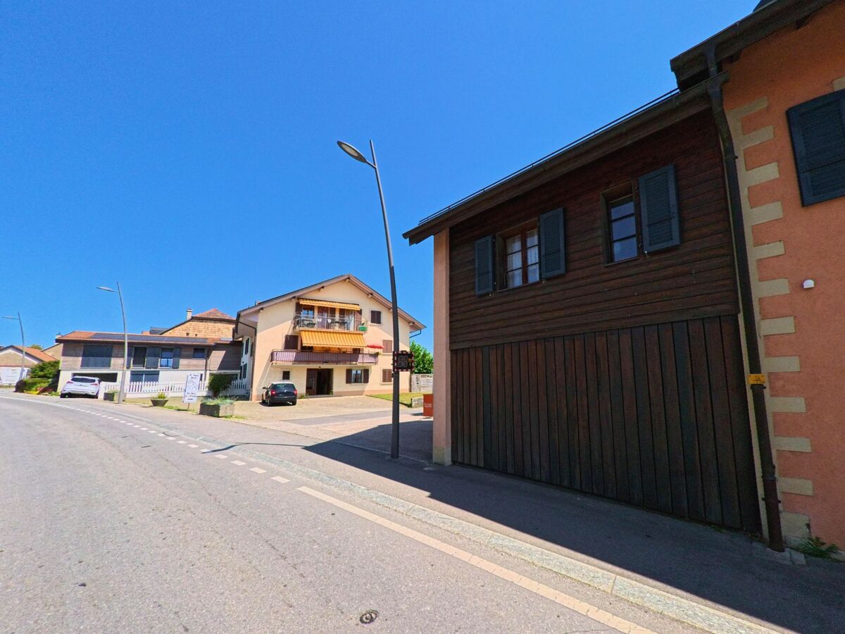 An einer leicht ansteigenden Strasse in Clarmont (1127, Kanton Waadt) stehen Wohnhäuser mit Balkonen und Garagen, daneben geparkte Autos unter klarem Himmel.