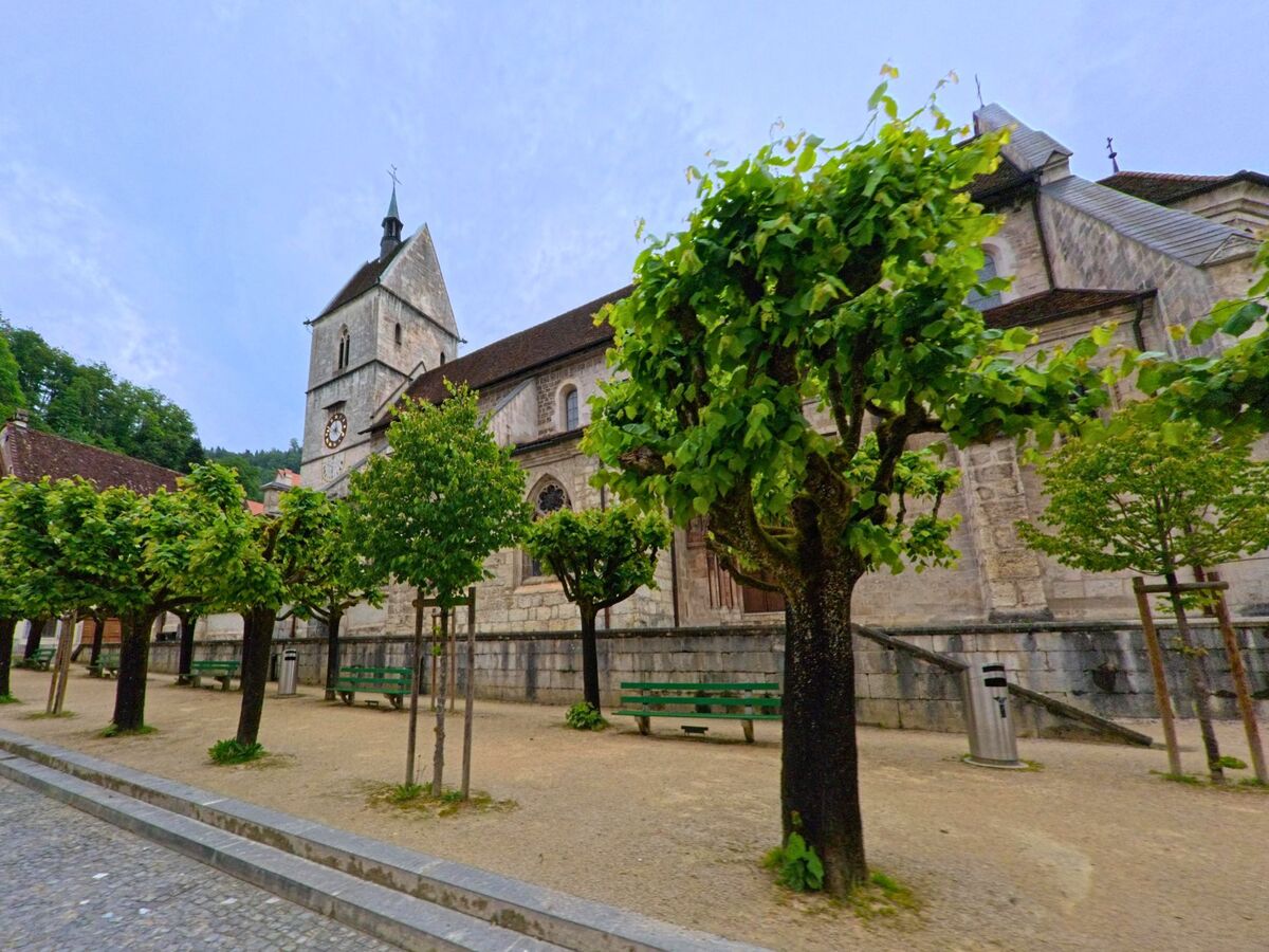 Vor der historischen Kirche in Clos du Doubs (2882, Kanton Jura) stehen in Reih und Glied geschnittene Bäume und grüne Bänke auf einem gepflasterten Platz.