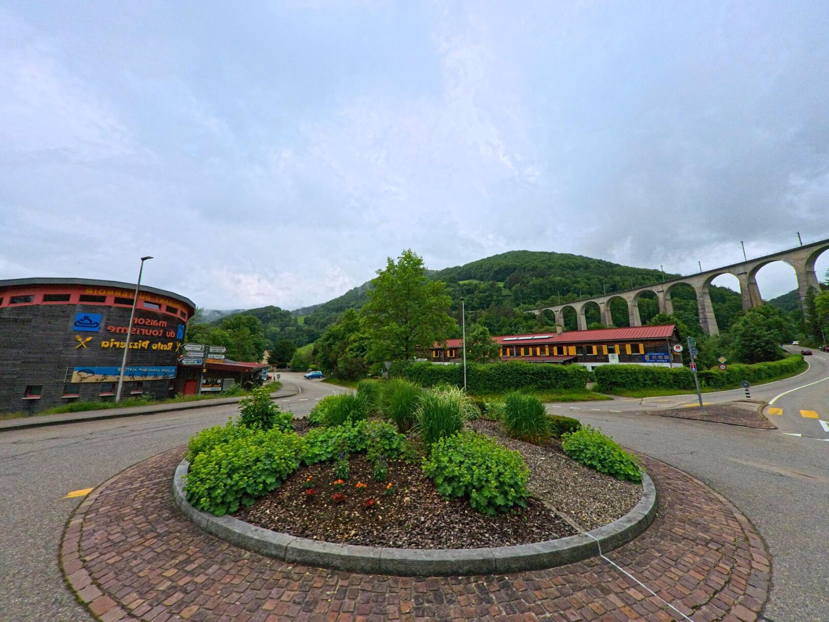In Clos du Doubs (2882, Kanton Jura) liegt ein begrünter Verkehrskreisel am Fuss eines Eisenbahnviadukts, umgeben von modernen Gebäuden und bewaldeten Hügeln.