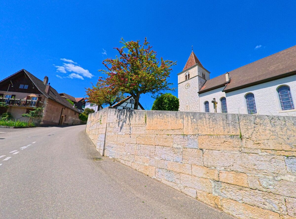 In Coeuve (PLZ 2932, Kanton Jura) fuehrt eine ansteigende Strasse an einer Kirche mit Glockenturm und einer Stützmauer entlang, begleitet von einem blühenden Baum.