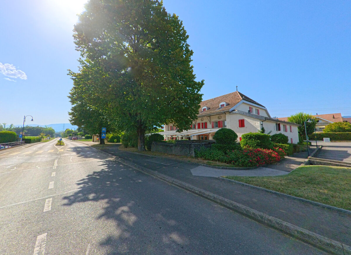 Ein traditionelles Haus mit roten Fensterlaeden steht in Coinsins (1267, Kanton Waadt) neben einer Hauptstrasse, die von Baeumen beschattet wird.