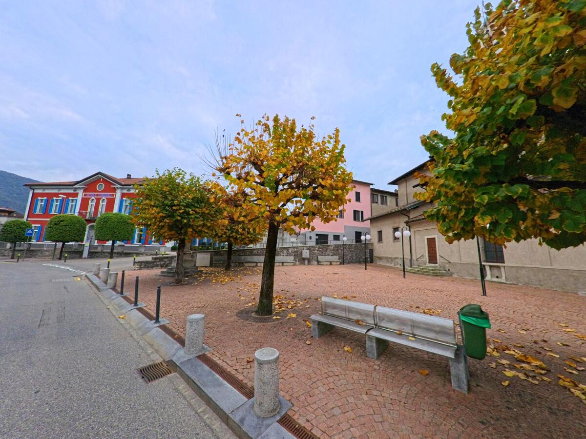 Ein kleiner Platz mit Bänken und herbstlich gefärbten Bäumen liegt im Zentrum von Coldrerio (6877, Kanton Tessin) vor dem roten Schulhaus.