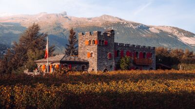 Das Schlossähnliche Gebäude der Colline de Daval in Sierre (3960, Kanton Wallis) liegt von Weinreben umgeben vor einer beeindruckenden Bergkulisse mit herbstlich gefärbter Landschaft.