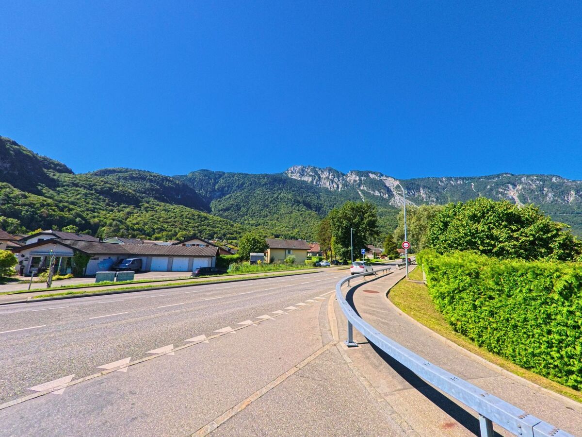 Eine breite Strasse fuehrt durch ein Wohngebiet von Collombey-Muraz (1893, Kanton Wallis) am Fusse eines bewaldeten Berghangs.